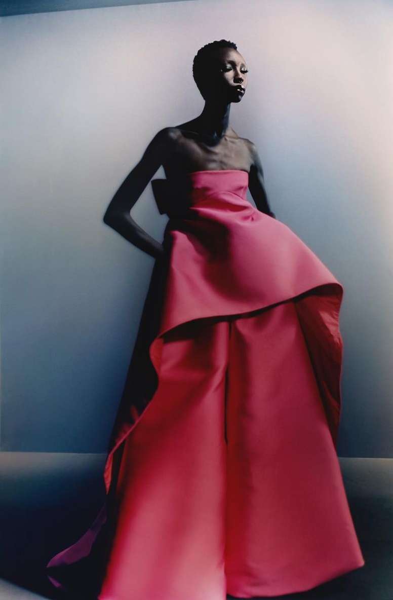 Fashion editorial image featuring a slender Black woman in an elegant, strapless, pink gown with a large bow, against a gradient background.