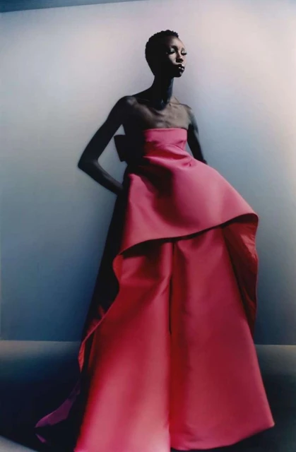 Fashion editorial image featuring a slender Black woman in an elegant, strapless, pink gown with a large bow, against a gradient background.