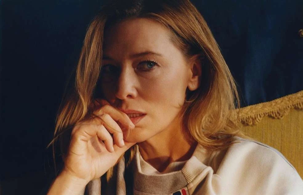 Close-up portrait of an adult woman with blonde hair, wearing a sweatshirt, looking at the camera while seated against a dark backdrop.
