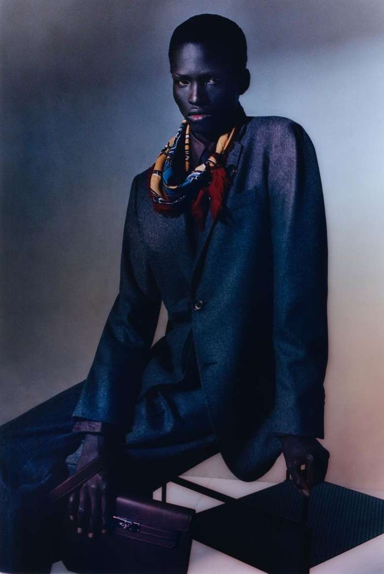 Fashion portrait of a Black model in an oversized suit and patterned silk scarf, seated with a leather purse, against a gradient backdrop.
