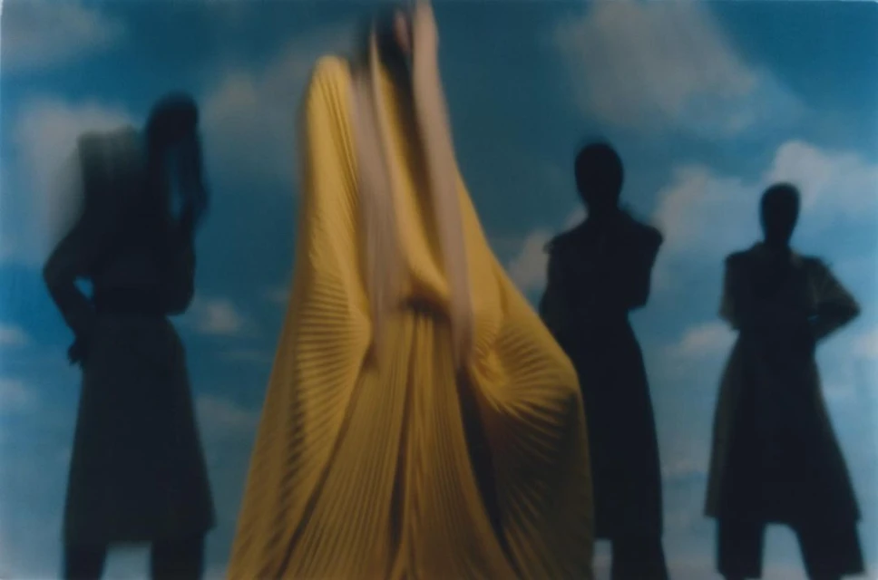 Fashion image with a model in a yellow pleated dress, silhouetted figures, against a backdrop of a blue sky with clouds creating a dreamy effect.