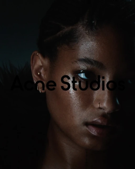 Close-up portrait of an African American woman with facial piercings, braided hair, and dark clothing, set against a dark background.