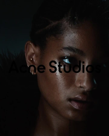 Close-up portrait of an African American woman with facial piercings, braided hair, and dark clothing, set against a dark background.
