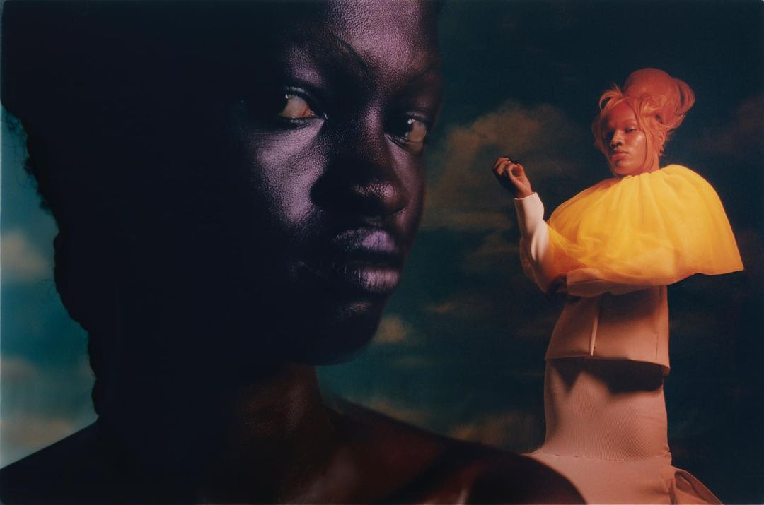 Striking image of two African women, one close-up with dark skin and one full-body with yellow tulle collar, against a cloudy background.