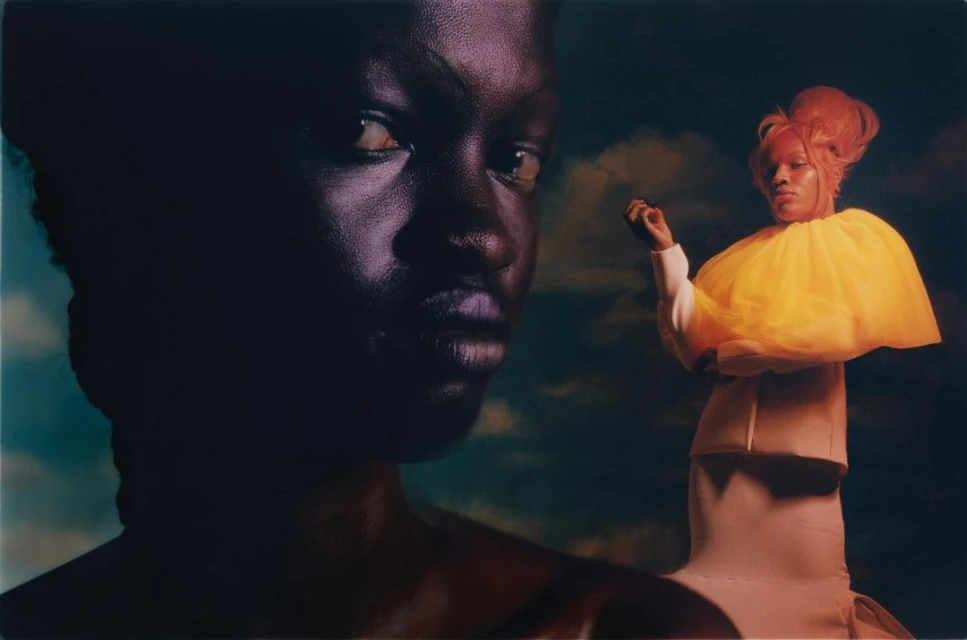 Striking image of two African women, one close-up with dark skin and one full-body with yellow tulle collar, against a cloudy background.
