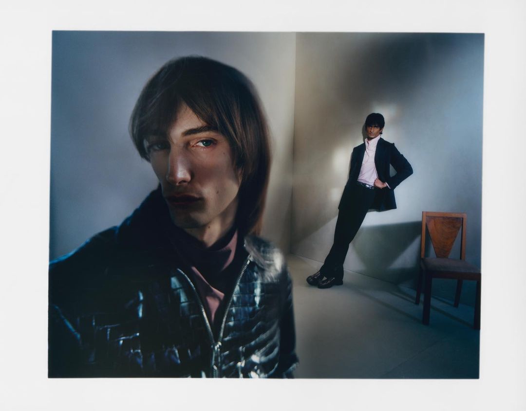 A stylish photograph features two young adult male models in suits, posed in an indoor setting with subdued lighting and a minimalist aesthetic.