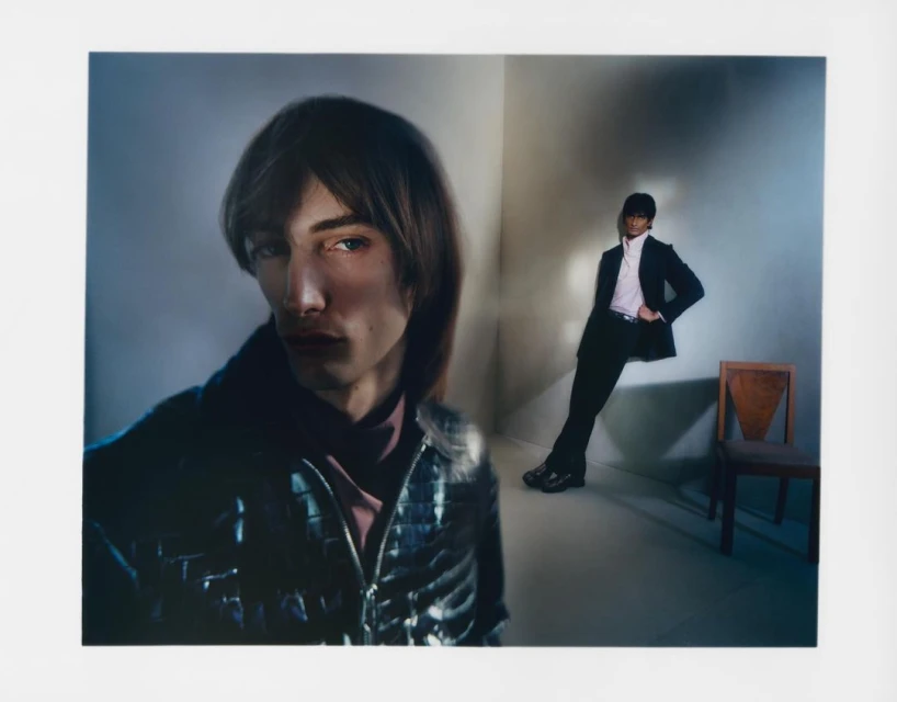 A stylish photograph features two young adult male models in suits, posed in an indoor setting with subdued lighting and a minimalist aesthetic.