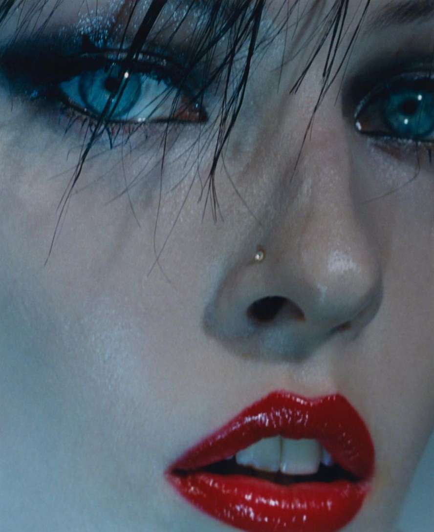 Close-up portrait of a woman with wet dark hair, piercing blue eyes, glossy red lipstick, and a tiny nose stud.