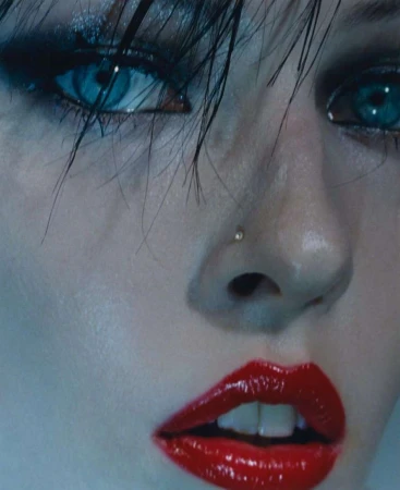Close-up portrait of a woman with wet dark hair, piercing blue eyes, glossy red lipstick, and a tiny nose stud.