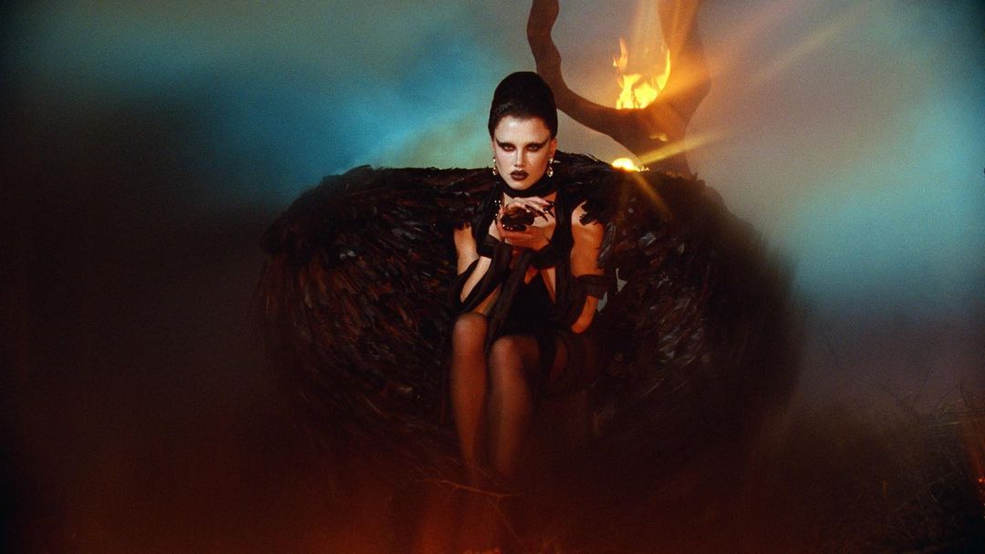 A woman in dark attire sits amidst a dark, smoky, and fiery scene, illuminated by orange and blue lighting, creating a dramatic atmosphere.