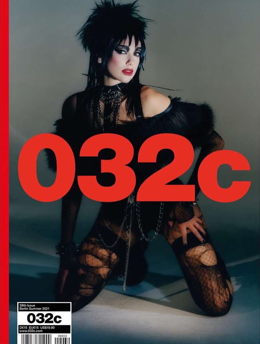 Magazine cover features a model with dark hair and makeup wearing a black outfit and fishnet stockings sitting in a dimly lit studio.
