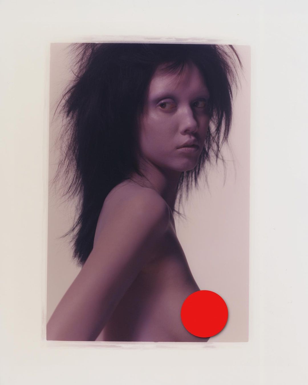 A portrait of a woman with dark, tousled hair, fair skin, and a semi-nude pose against a neutral backdrop, with a red circle obscuring the chest.