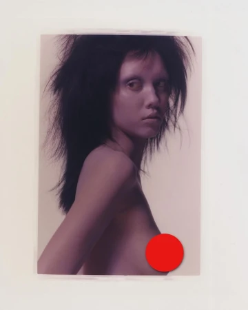 A portrait of a woman with dark, tousled hair, fair skin, and a semi-nude pose against a neutral backdrop, with a red circle obscuring the chest.