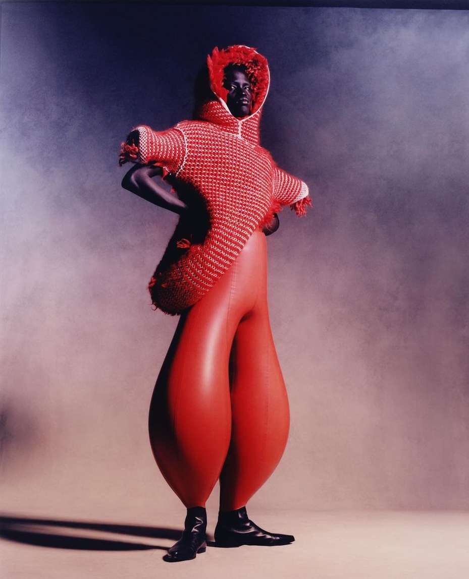 Runway fashion features an African American model in a red and white knitted hooded top with bulbous red trousers against a smoky studio backdrop.