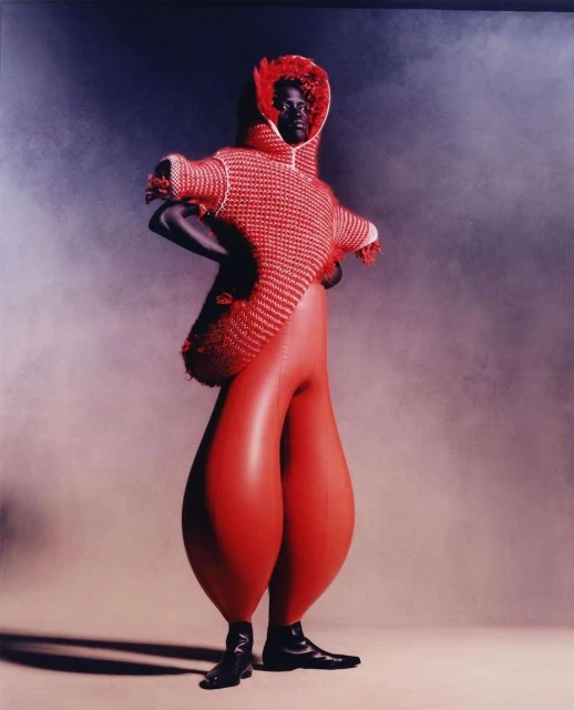 Runway fashion features an African American model in a red and white knitted hooded top with bulbous red trousers against a smoky studio backdrop.