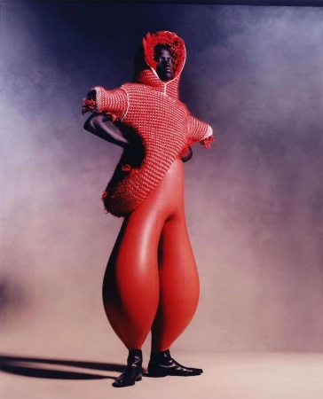 Runway fashion features an African American model in a red and white knitted hooded top with bulbous red trousers against a smoky studio backdrop.