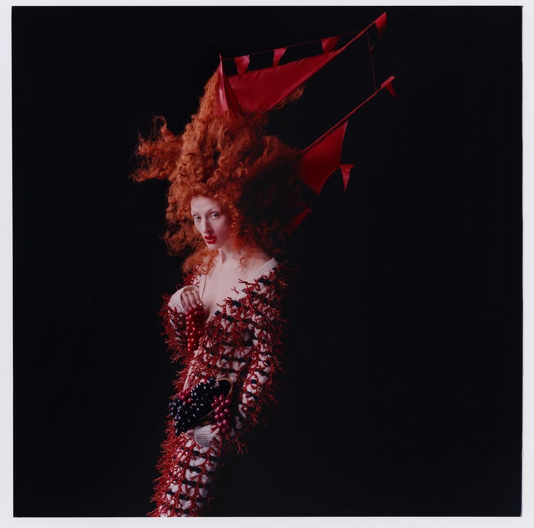 Elegant woman with red hair and a decorative dress, posing against a dark background in a studio fashion editorial shot.