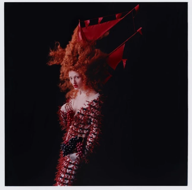 Elegant woman with red hair and a decorative dress, posing against a dark background in a studio fashion editorial shot.