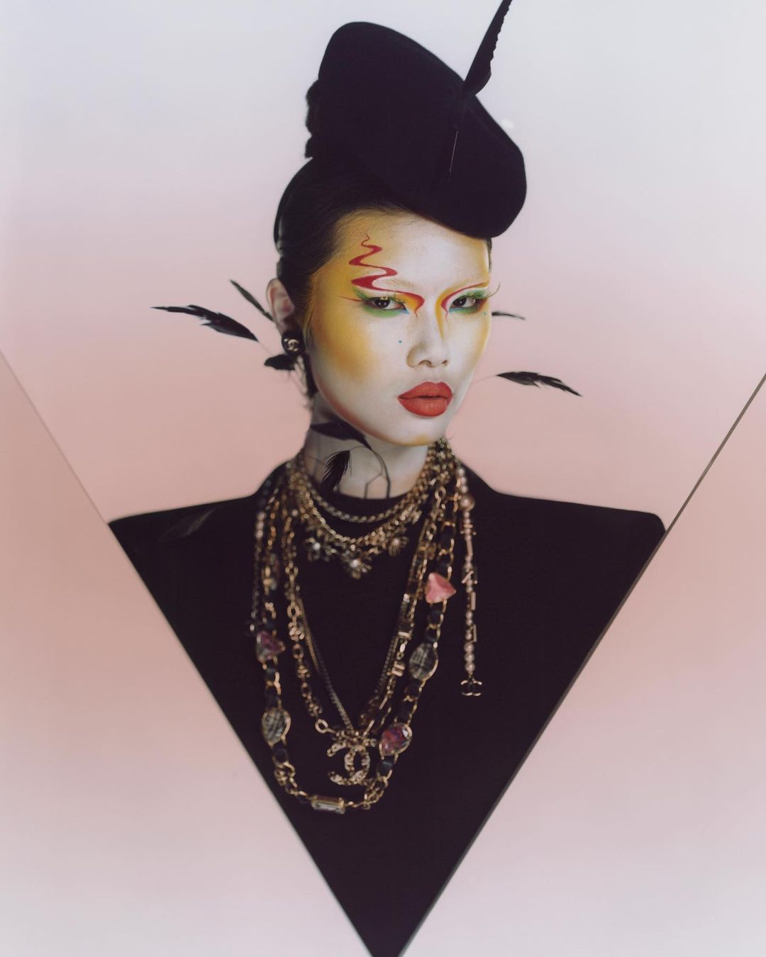 A person with a striking makeup look, wearing a black hat and multiple necklaces, poses against a pink and black backdrop.