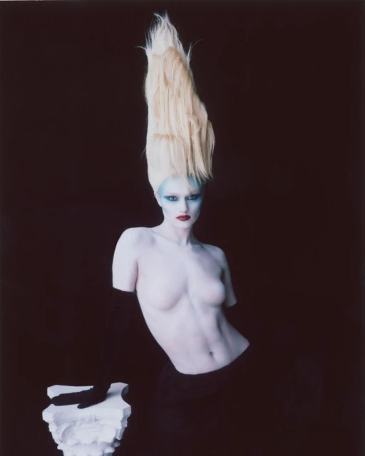 Dramatic portrait of a pale woman with striking makeup and towering blonde hair, posing with a sculpted pedestal against a dark backdrop.