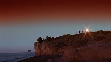 A rocky cliff landscape meets the sea under an orange-to-blue gradient sky. A group of people stand at the cliff's edge, silhouetted against a bright glare.