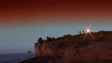 A rocky cliff landscape meets the sea under an orange-to-blue gradient sky. A group of people stand at the cliff's edge, silhouetted against a bright glare.