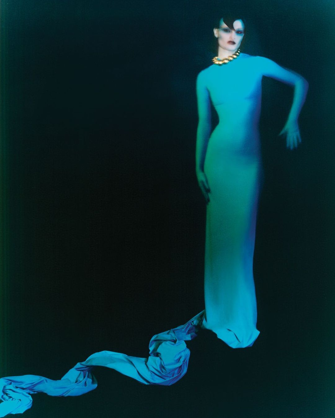 Elegant woman in a long, body-conscious dress and bold gold necklace poses against a deep backdrop in a dramatic fashion shot.