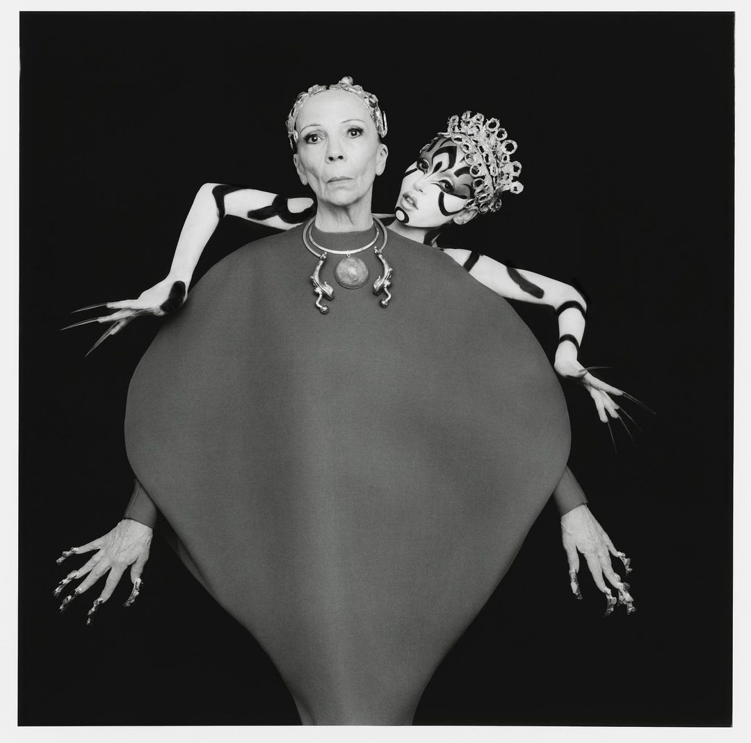 Two women posing together against a black background wearing avant-garde headpieces and elaborate makeup.