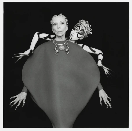 Two women posing together against a black background wearing avant-garde headpieces and elaborate makeup.