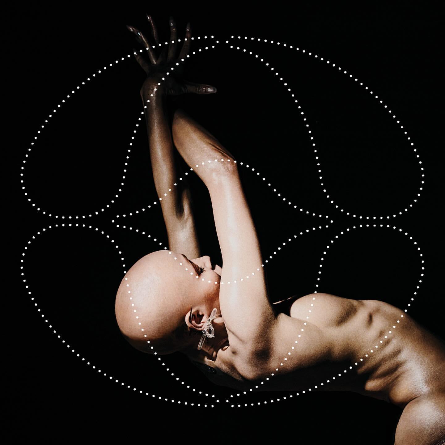 Dramatic studio shot of a bald woman in dynamic pose, arms raised, body arched, adorned with geometric dot pattern against a black background.