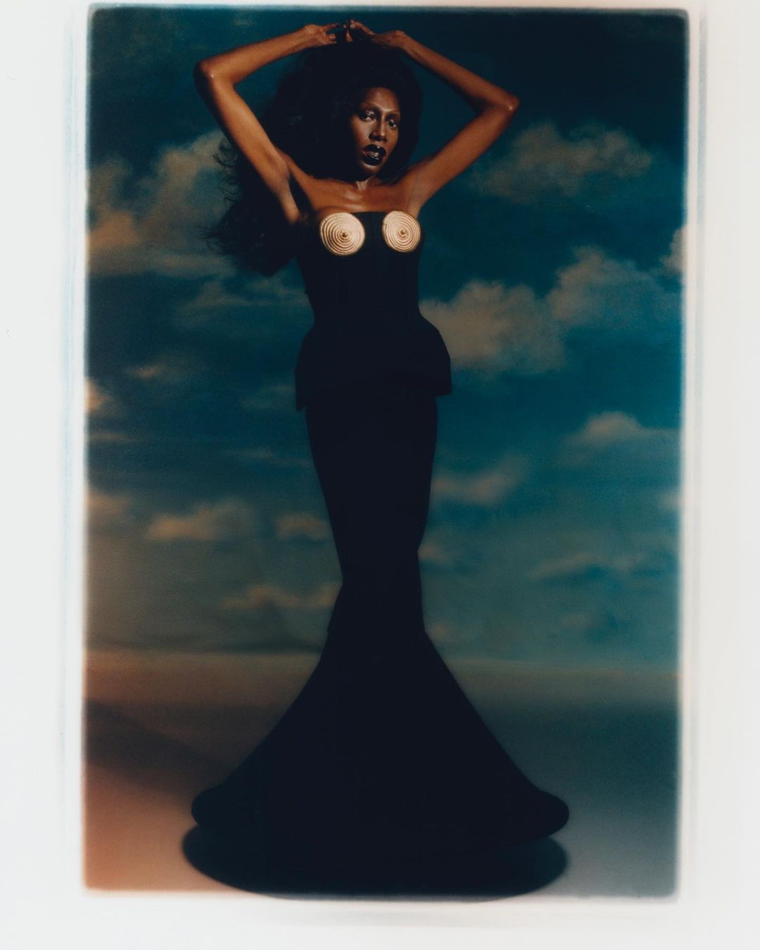 Glamour shot of a slender African-American woman with long black hair in a long black dress against a sky and cloud backdrop.
