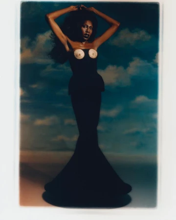 Glamour shot of a slender African-American woman with long black hair in a long black dress against a sky and cloud backdrop.