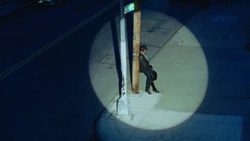 Aerial shot of adult male leaning against a light post in an urban setting with dark blue tones and harsh spotlight effect at night.