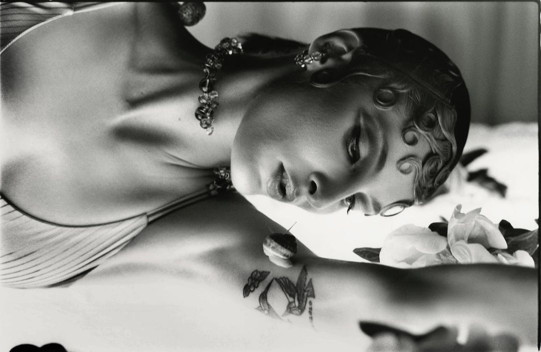 Black and white portrait of a woman with sculpted baby hair, beaded necklace, and a small snail on her tattooed arm by Jordan Hemingway