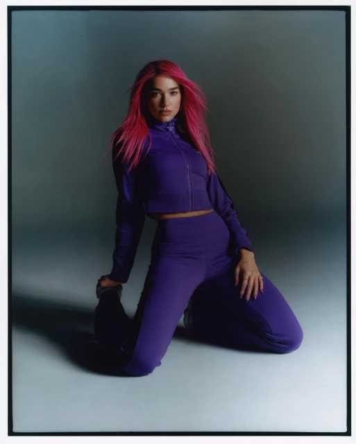 Portrait of an adult woman with magenta hair, posing while kneeling in a matching purple tracksuit against a muted grey background.