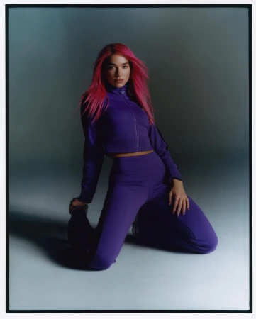 Portrait of an adult woman with magenta hair, posing while kneeling in a matching purple tracksuit against a muted grey background.
