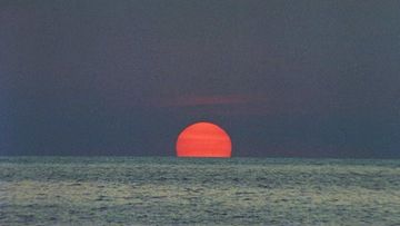 Dramatic sunset over the ocean, featuring a vivid orange sun partially submerged on the water horizon, creating a peaceful and serene scene.