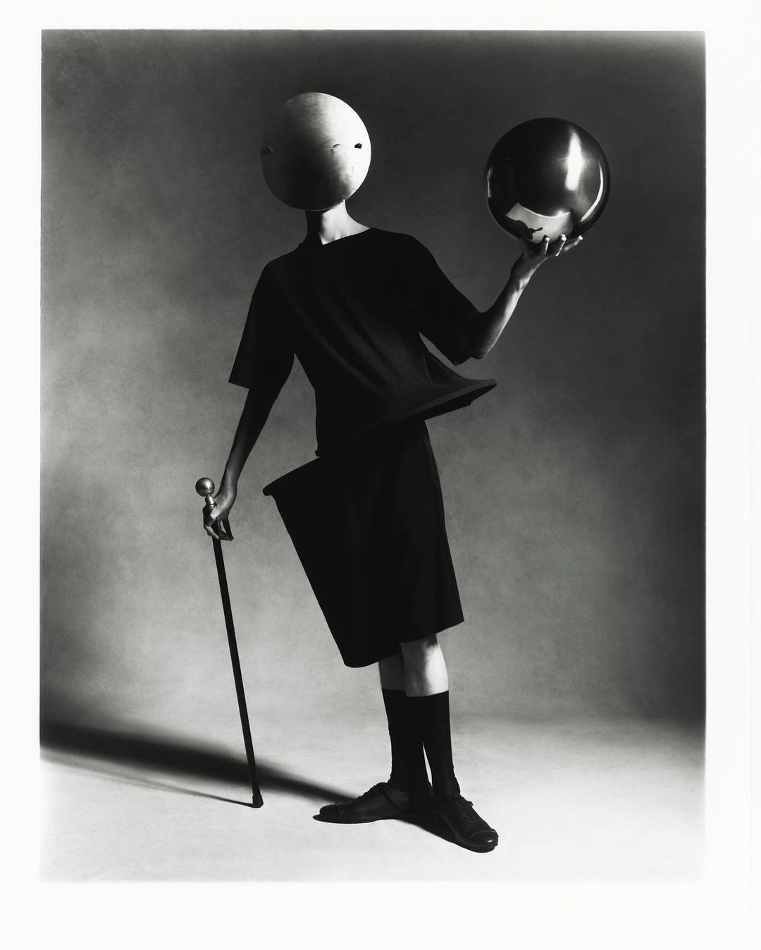 A person wearing black clothing and a spherical mask balances a shiny ball on their hand, holding a cane, in a studio setting with soft lighting.