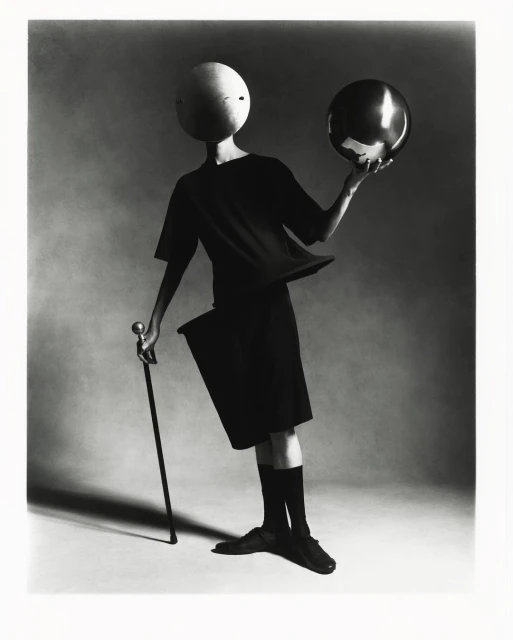 A person wearing black clothing and a spherical mask balances a shiny ball on their hand, holding a cane, in a studio setting with soft lighting.