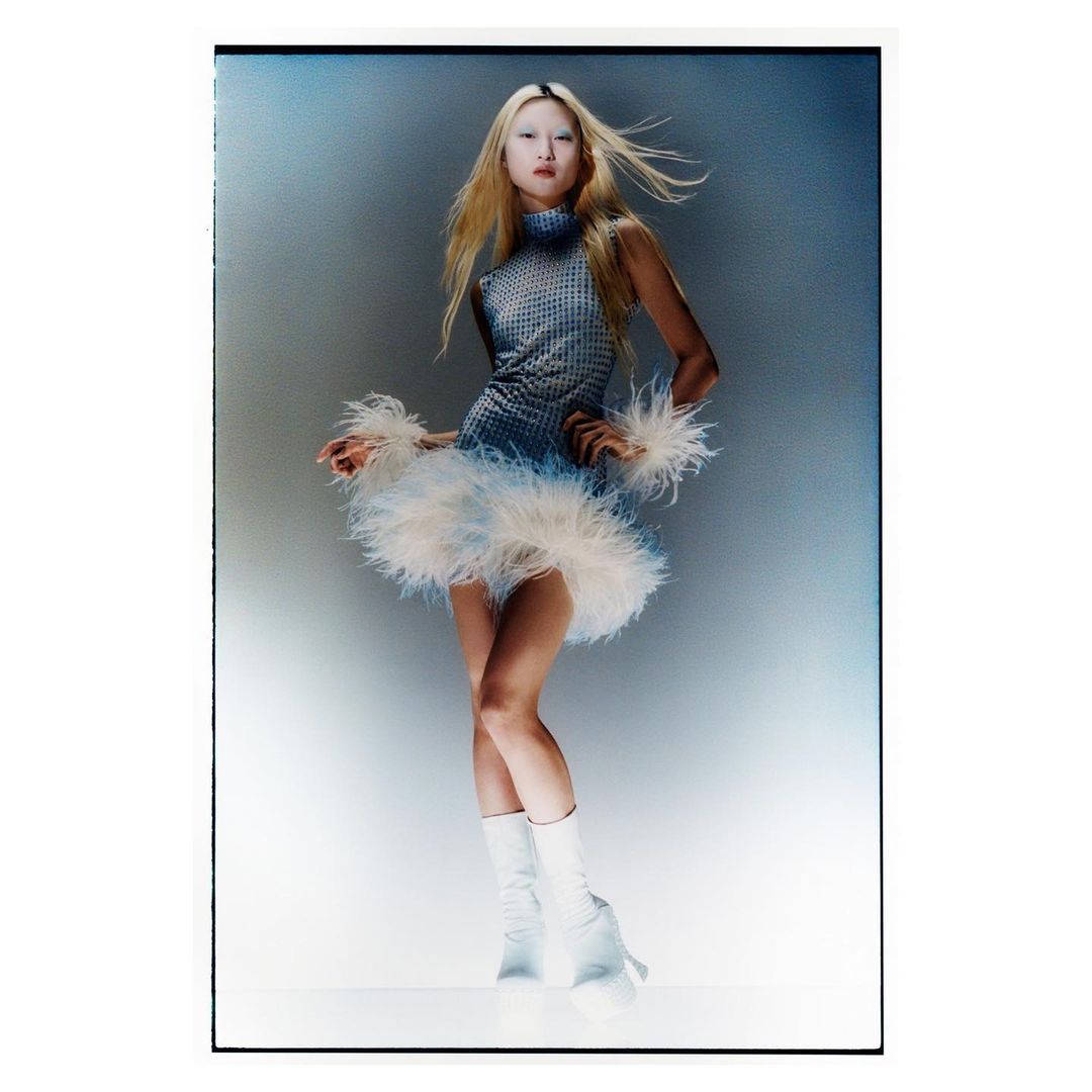 Full-length portrait of a young woman with blonde hair, wearing a blue mini dress with feathered hem and white platform boots in studio lighting.