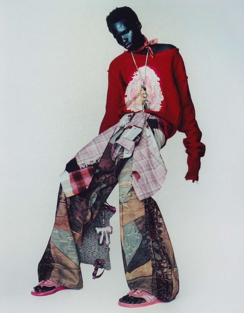 Fashionable person posing in oversized patchwork pants, red sweater, neck scarf, and pink platform shoes against a plain white background.