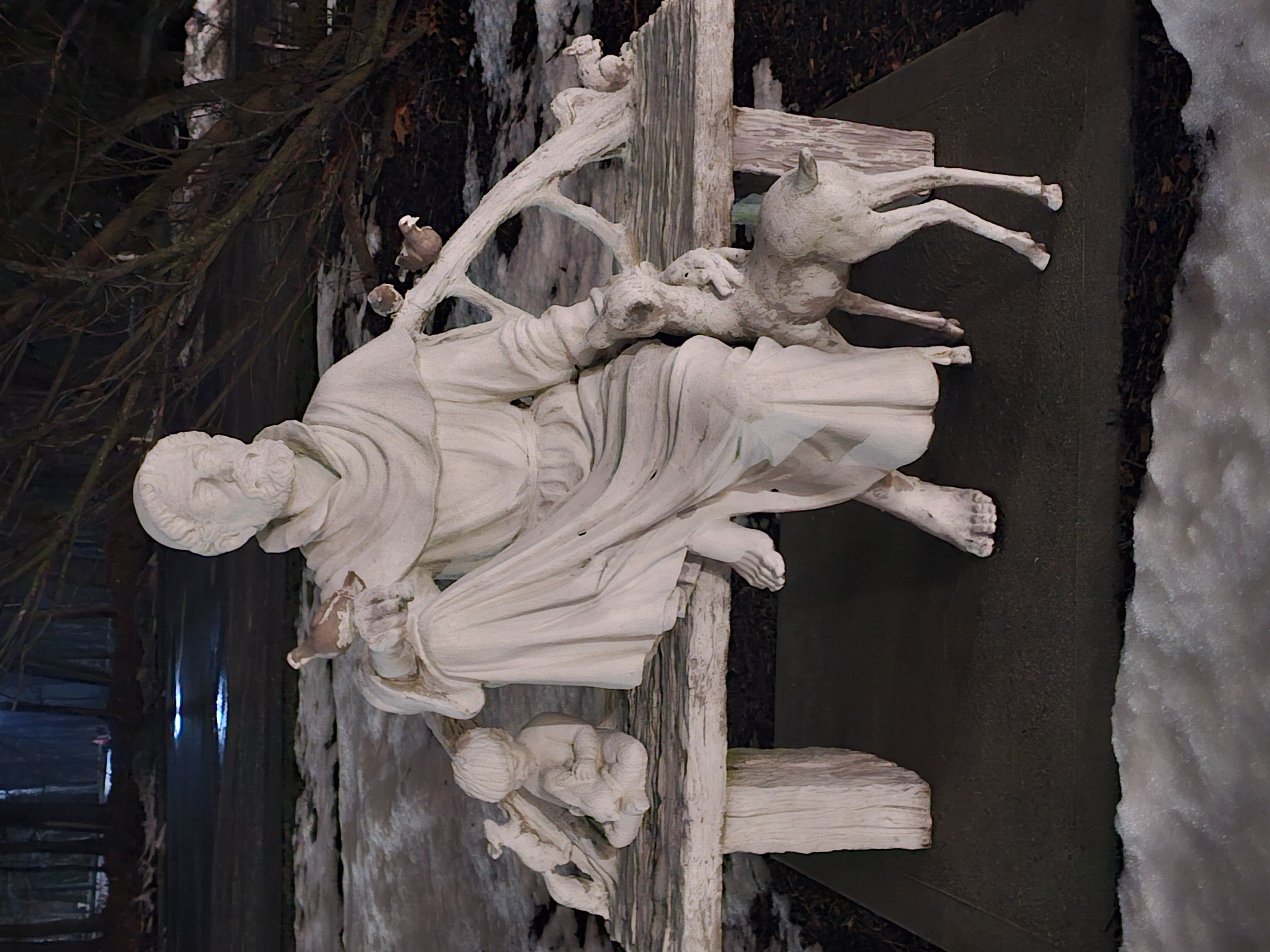 Marble statue featuring a seated robed man surrounded by a fawn, child, and birds in a snowy park setting at dusk or dawn.
