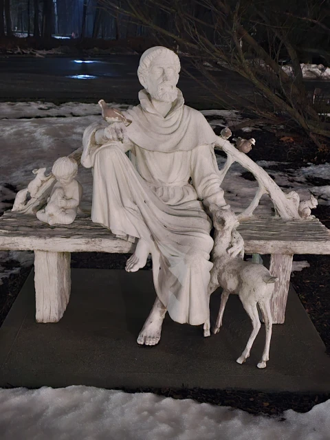 Marble statue featuring a seated robed man surrounded by a fawn, child, and birds in a snowy park setting at dusk or dawn.