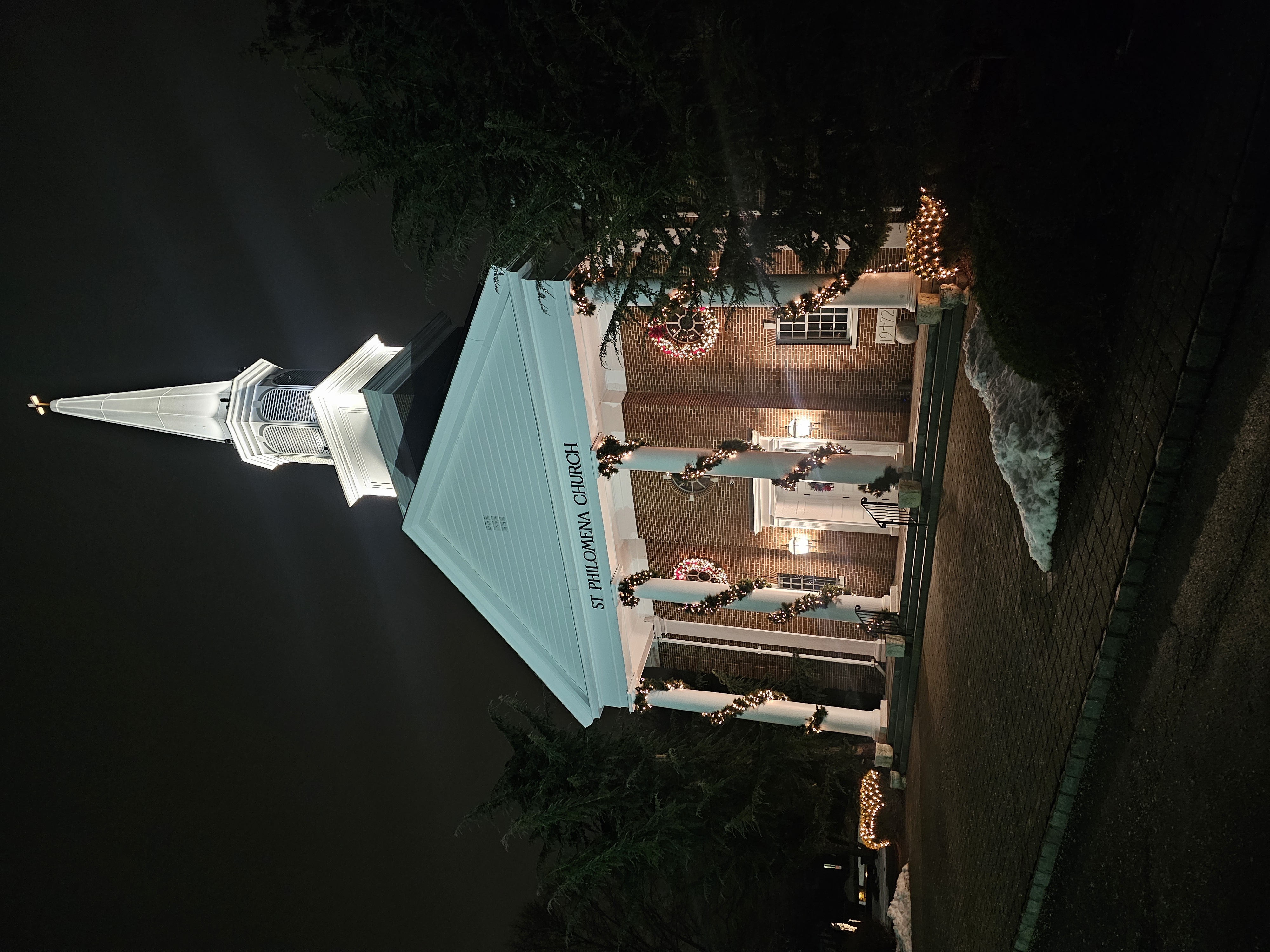 Exterior view of the Saint Philomena Church at night, adorned with Christmas wreaths and garland, showcasing architectural details and festive lighting.