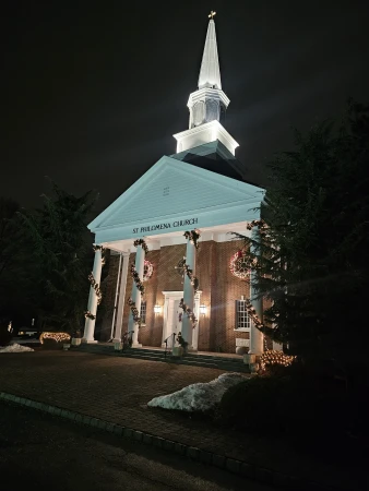Exterior view of the Saint Philomena Church at night, adorned with Christmas wreaths and garland, showcasing architectural details and festive lighting.