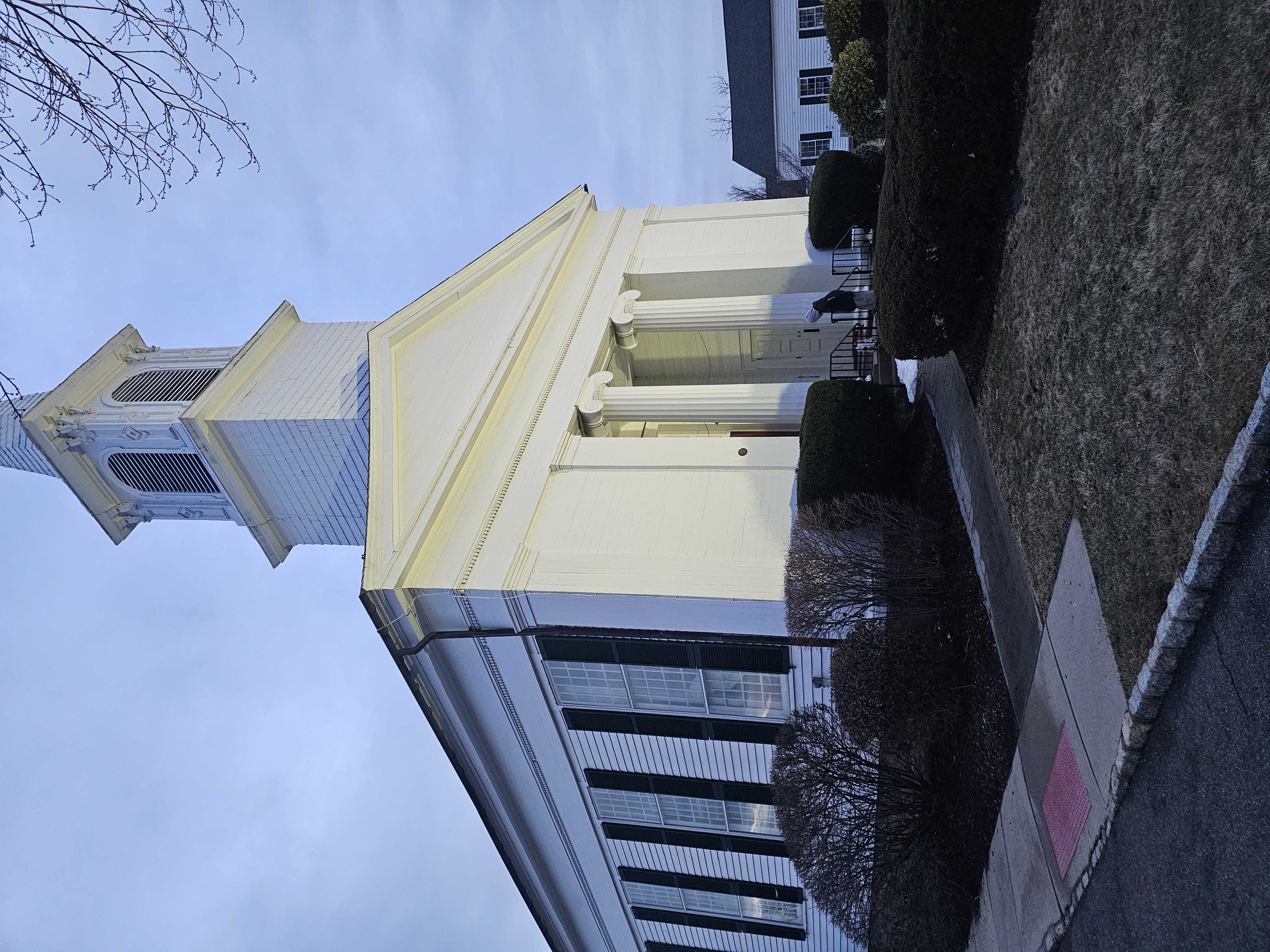 White church building with a steeple, framed by bushes and trees under a blue sky, capturing architectural details and serene ambiance.
