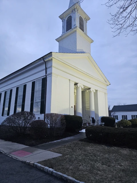 White church building with a steeple, framed by bushes and trees under a blue sky, capturing architectural details and serene ambiance.