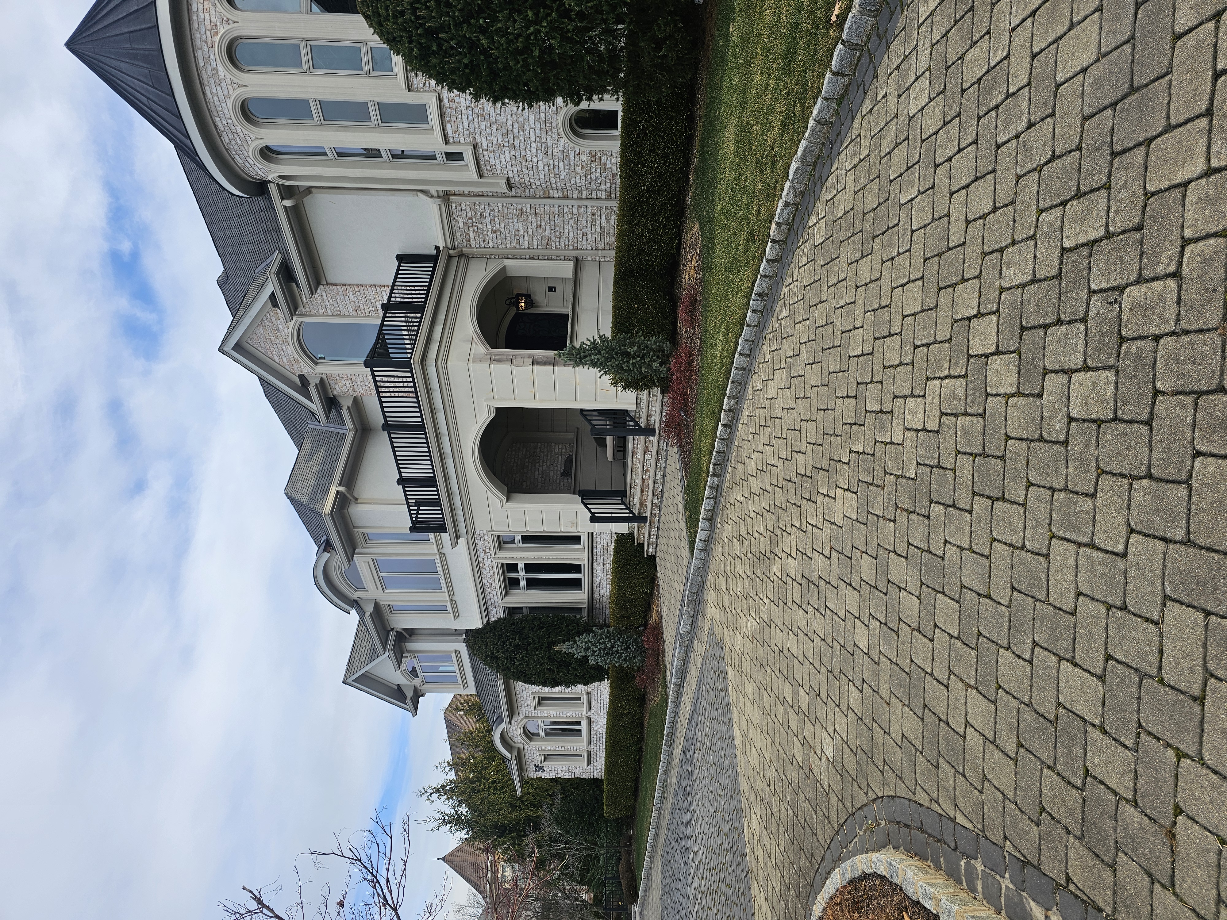 Exterior view of a large two-story residential house with a brick driveway, manicured hedges, and a cloudy sky on a sunny day.