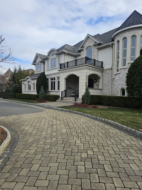Exterior view of a large two-story residential house with a brick driveway, manicured hedges, and a cloudy sky on a sunny day.