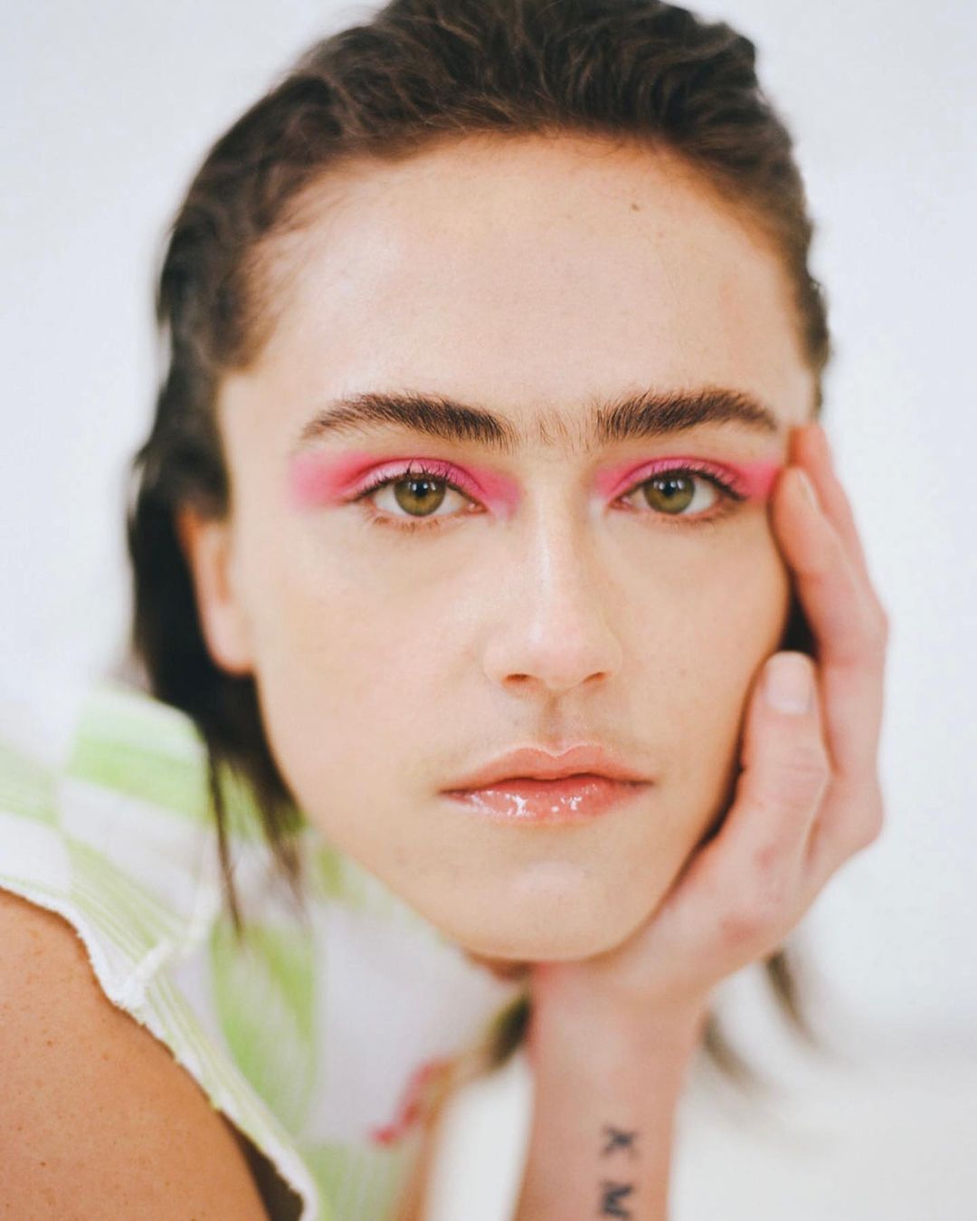 Close-up portrait of a person with pink eyeshadow, slicked-back dark hair, and a green and white shirt against a white background.